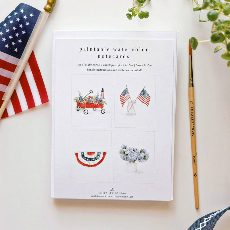 White notecard box displaying four Americana-themed watercolor illustrations: flower wagon, crossed flags in vase, red bunting, and hydrangea