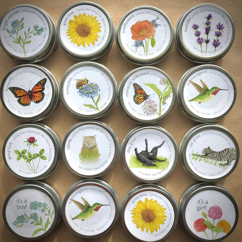 Grid of 16 seed packets in metal tins featuring botanical watercolor illustrations: flowers, butterflies, hummingbird, cat, owl, and birds on cream labels with gold rims.