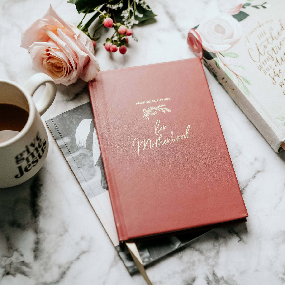 Dusty rose hardcover journal titled "Praying Scripture for Motherhood" with white text and leaf design, arranged on marble with peach rose, greenery, and coffee mug.