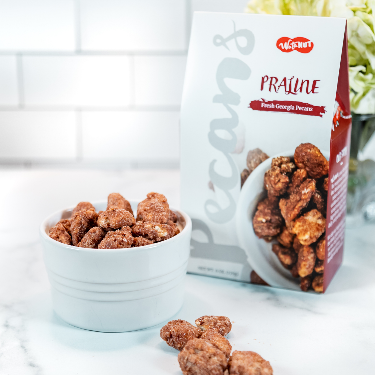 Crispy praline pecans in white ridged bowl with Watanut package, scattered nuts on marble surface