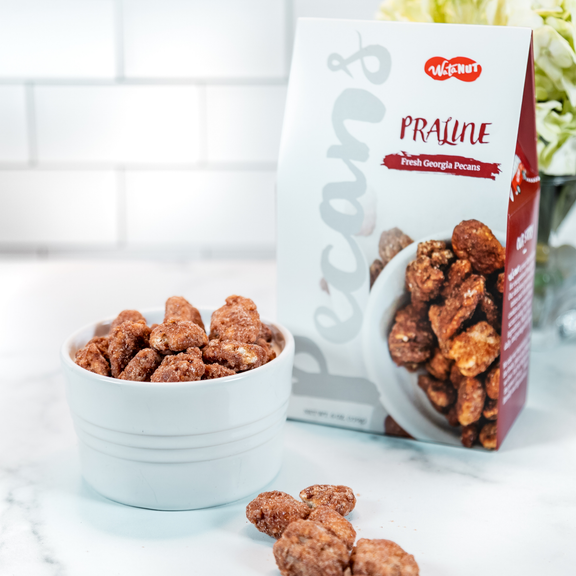 Crispy praline pecans in white ridged bowl with Watanut package, scattered nuts on marble surface