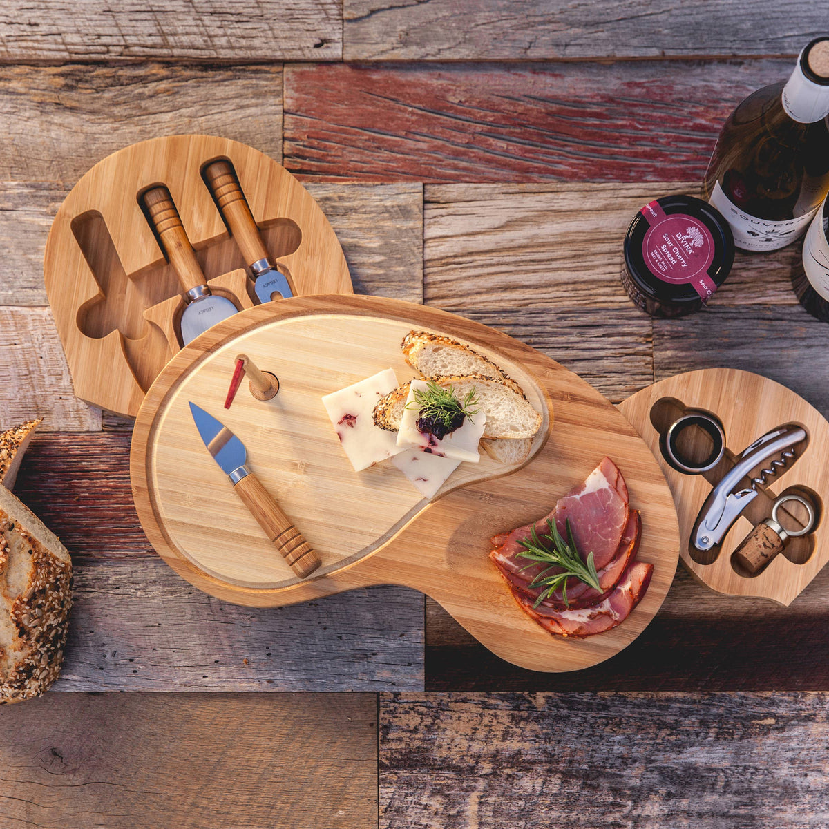 Bamboo golf-club-shaped cheese board featuring two connected cutting surfaces with cheese, charcuterie, stainless steel serving tools, and wine bottle.