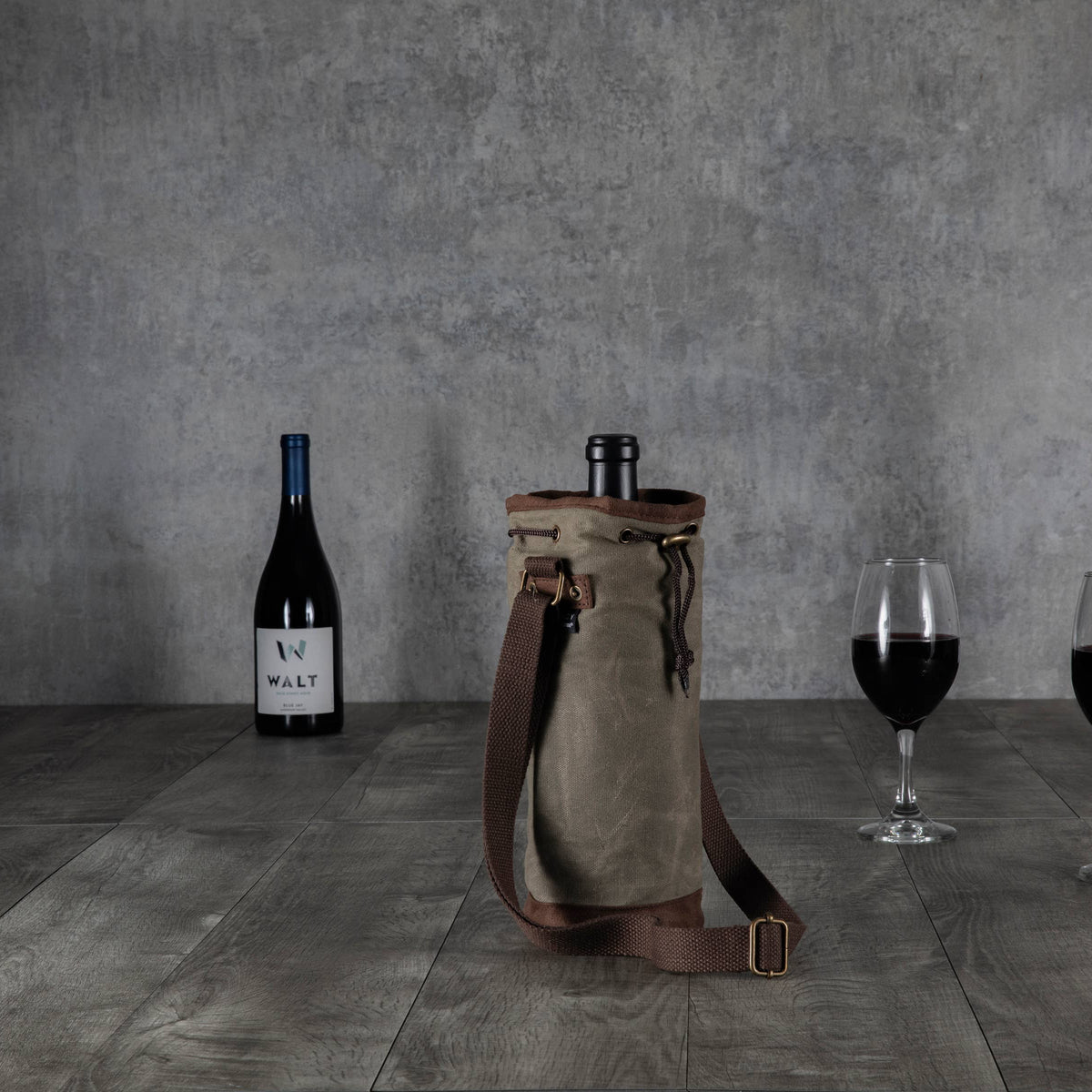 Tan waxed canvas wine tote with brown leather shoulder strap and metal hardware, displayed on dark wood flooring with wine bottles and glasses