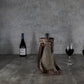 Tan waxed canvas wine tote with brown leather shoulder strap and metal hardware, displayed on dark wood flooring with wine bottles and glasses