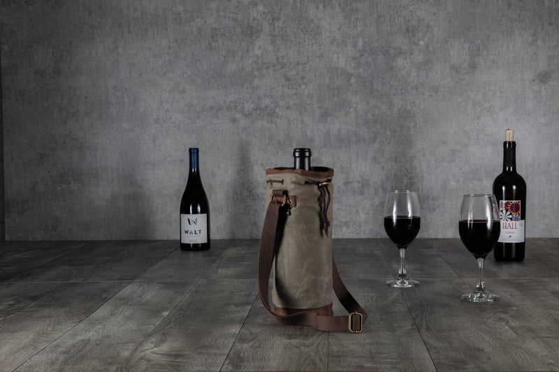 Tan waxed canvas wine tote with brown leather shoulder strap and metal hardware, displayed on dark wood flooring with wine bottles and glasses