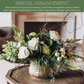 White roses, yellow ranunculus, fern fronds, and trailing branches in a clear glass vase on a wooden surface