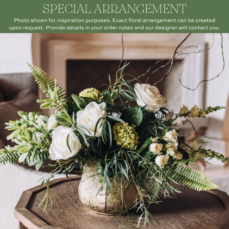 White roses, yellow ranunculus, fern fronds, and trailing branches in a clear glass vase on a wooden surface