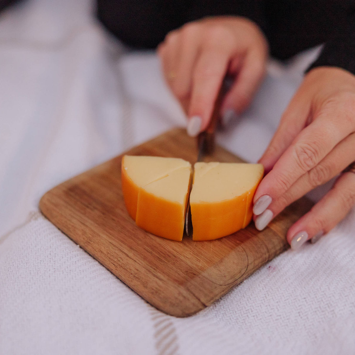 Knife cutting orange cheddar cheese into wedges on a wooden cutting board held in hands with black and white sleeves visible