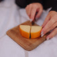 Knife cutting orange cheddar cheese into wedges on a wooden cutting board held in hands with black and white sleeves visible