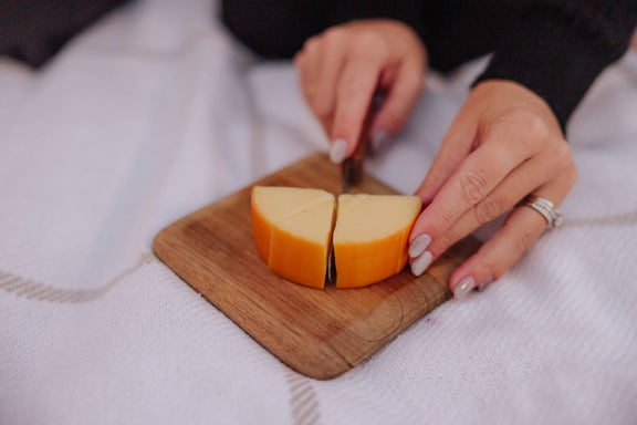 Knife cutting orange cheddar cheese into wedges on a wooden cutting board held in hands with black and white sleeves visible