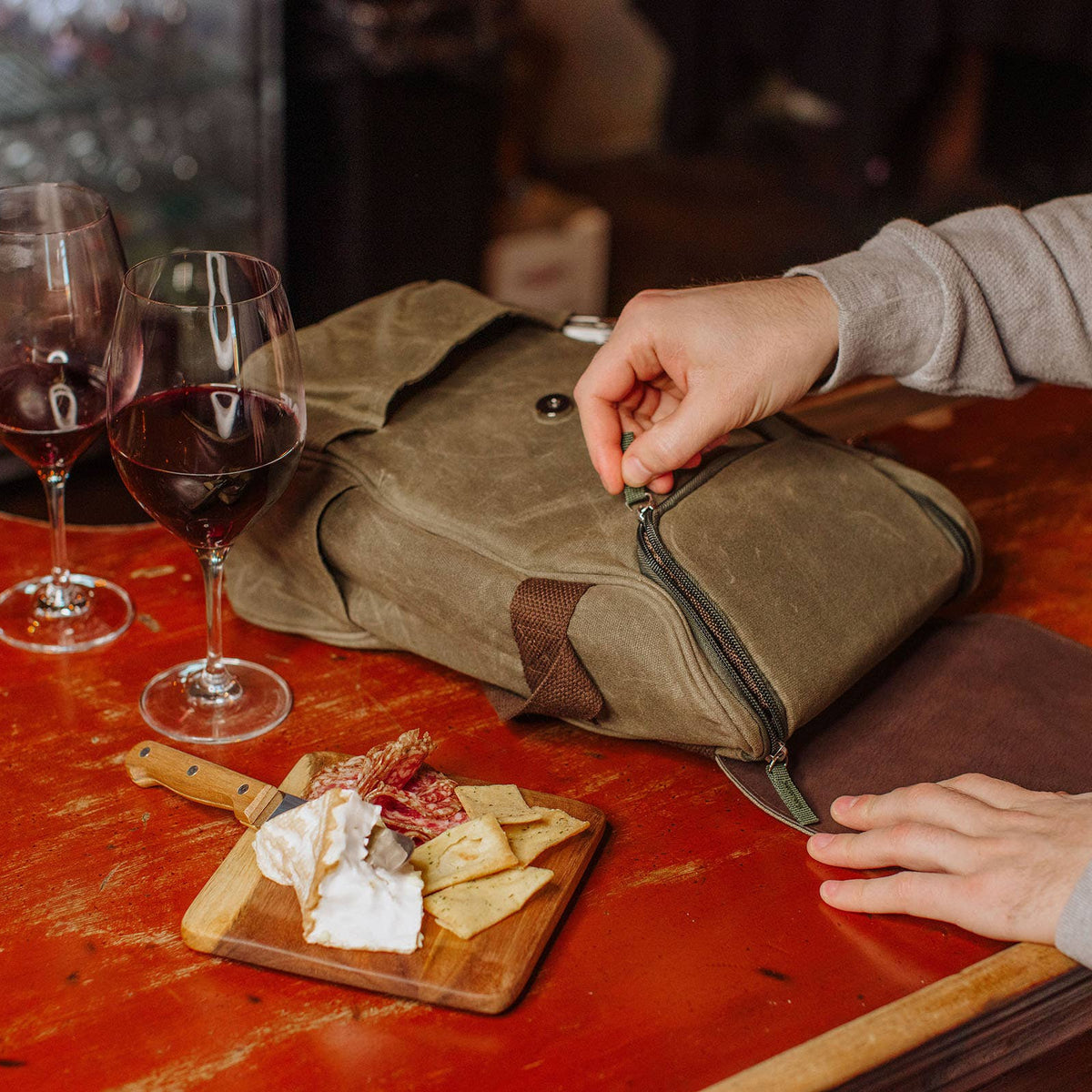 Olive-green insulated wine cooler bag opened on red table with two filled wine glasses and wooden cheese board with crackers and cheese