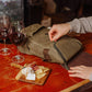 Olive-green insulated wine cooler bag opened on red table with two filled wine glasses and wooden cheese board with crackers and cheese