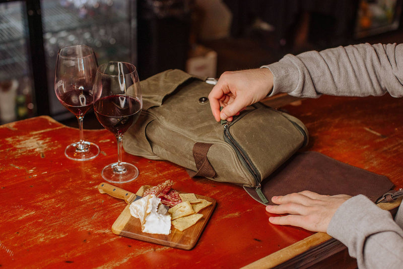 Olive-green insulated wine cooler bag opened on red table with two filled wine glasses and wooden cheese board with crackers and cheese