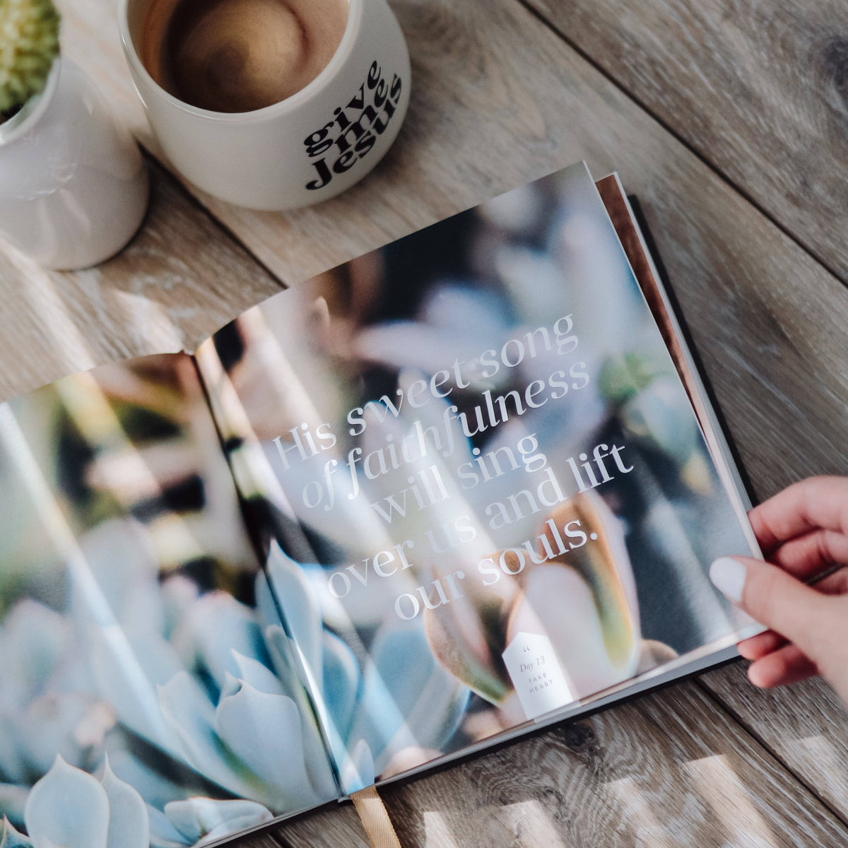 Open devotional book held in hand on wooden table beside white mug of coffee and potted succulent plant
