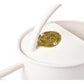 Buttermilk-colored indoor watering can with curved spout and handle, featuring a brass maker's plate on the rounded body