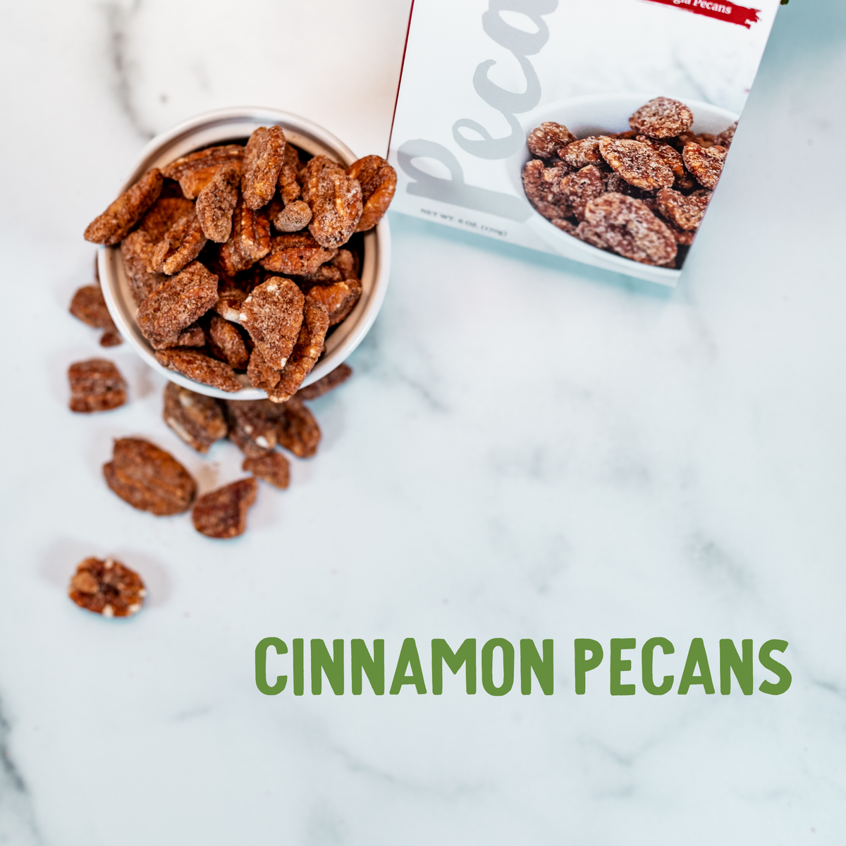 Roasted cinnamon pecans scattered from a white bowl on marble surface with Georgia State Gift Tin box.