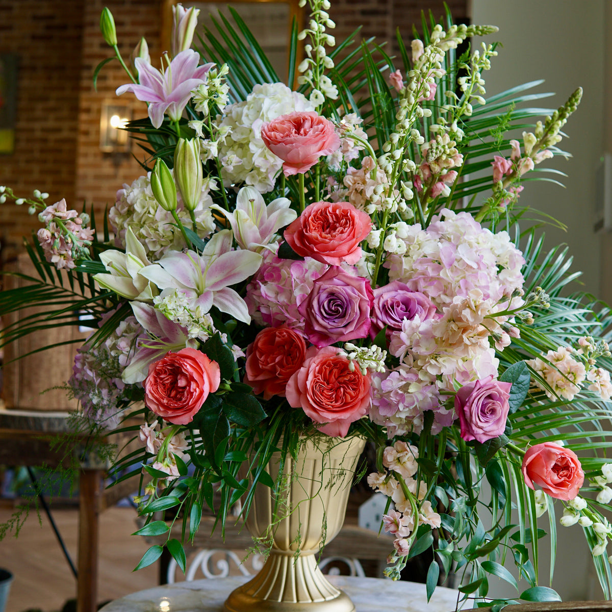 Tall sympathy arrangement in cream pedestal vase with red and pink roses, purple blooms, white lilies, light pink peonies, fern and green foliage on dark wood surface.