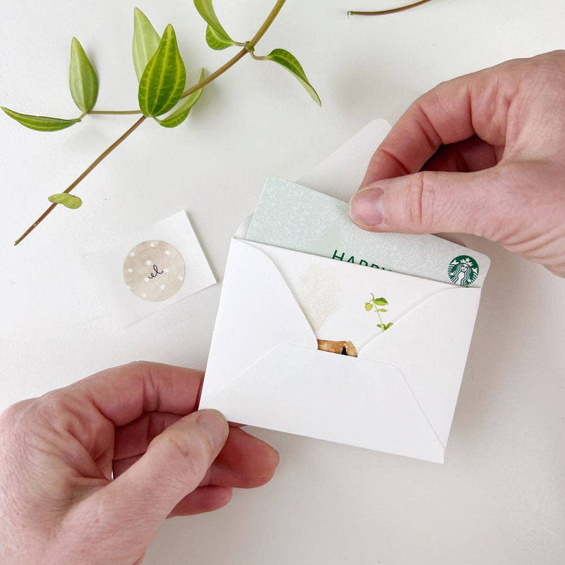 Two hands holding white notecards with light green mini card on top, envelope below, small "el" sticker, and green branch accent against white background