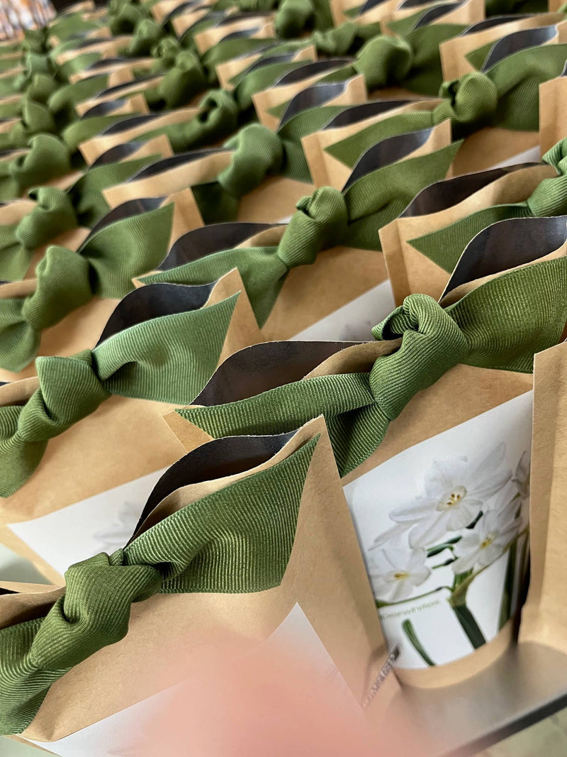 Rows of kraft paper bags tied with sage green ribbon bows, each containing paperwhite bulbs ready to plant