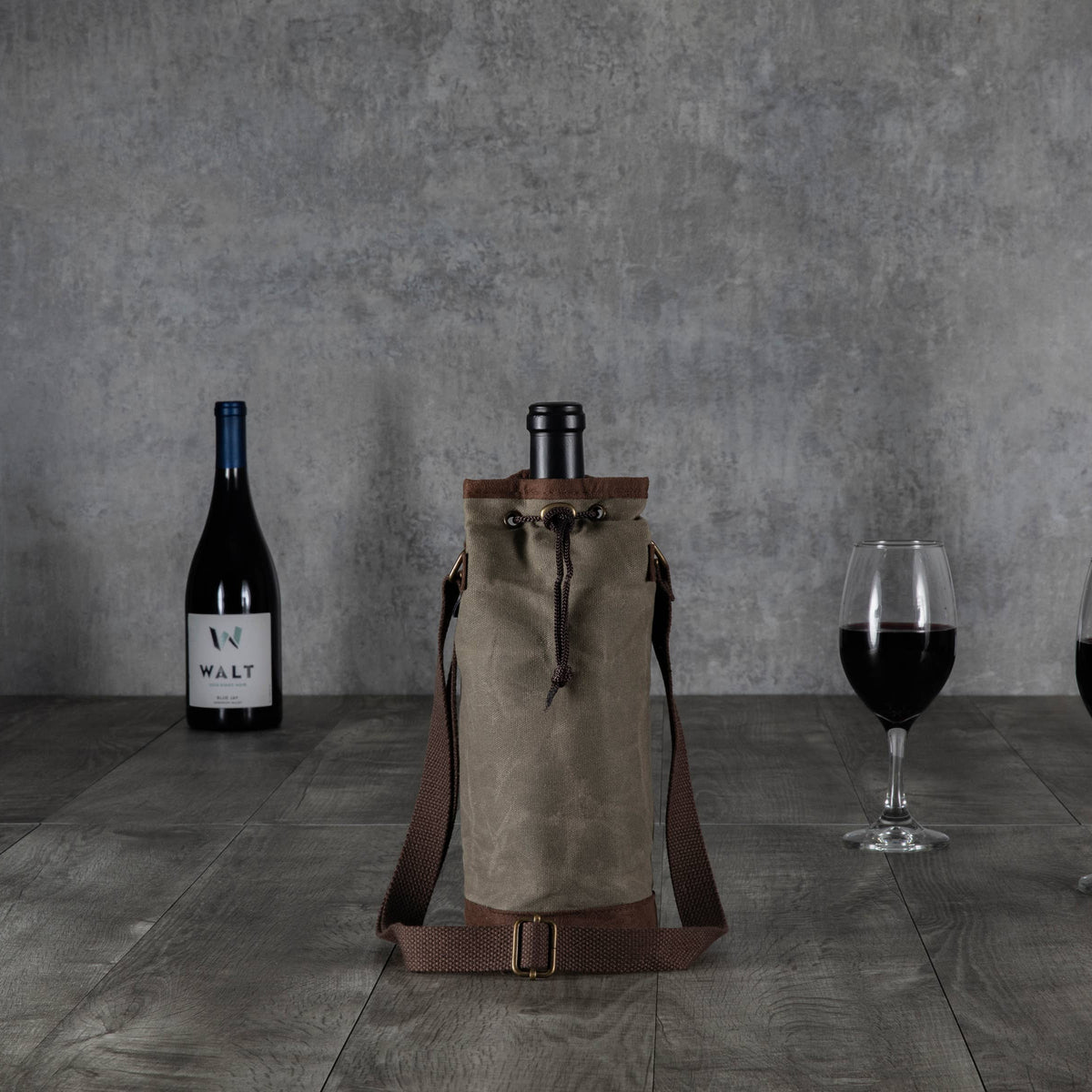 Beige waxed canvas wine tote with brown leather trim and adjustable shoulder straps, displayed with wine bottles and glasses