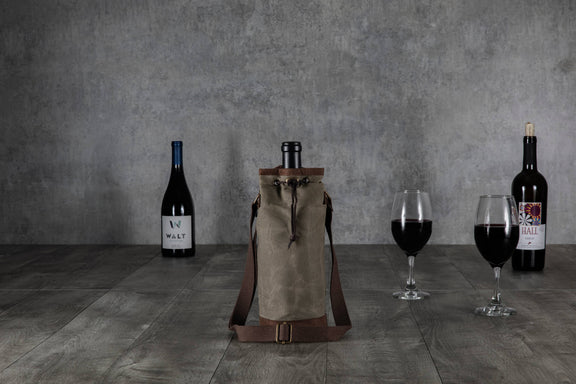 Beige waxed canvas wine tote with brown leather trim and adjustable shoulder straps, displayed with wine bottles and glasses