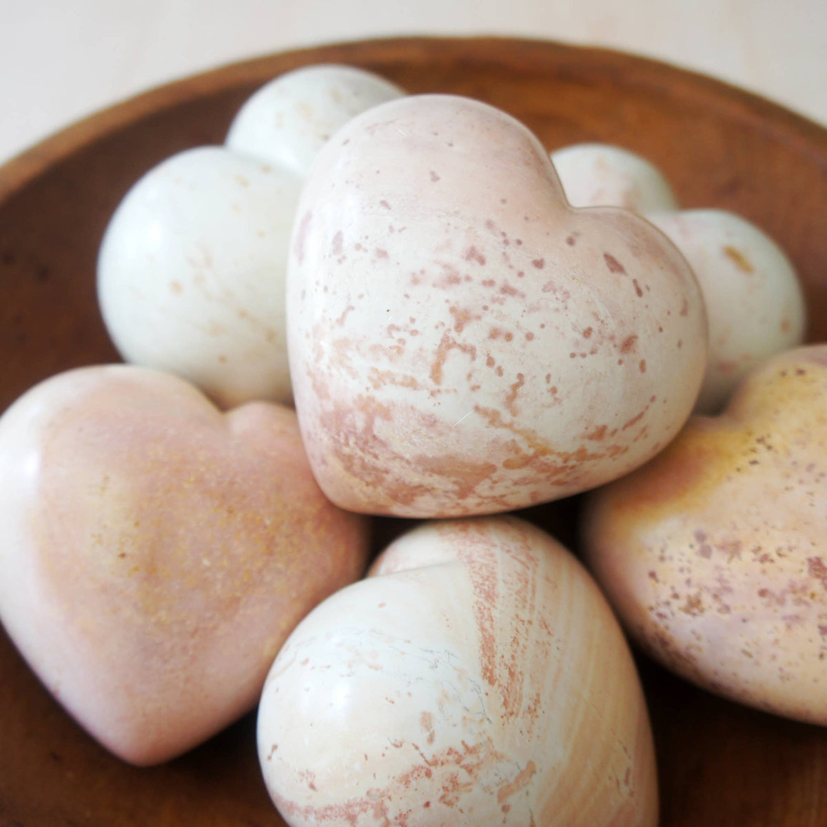 Hand-carved soapstone hearts in cream and blush tones with natural stone texture, arranged in a wooden bowl.