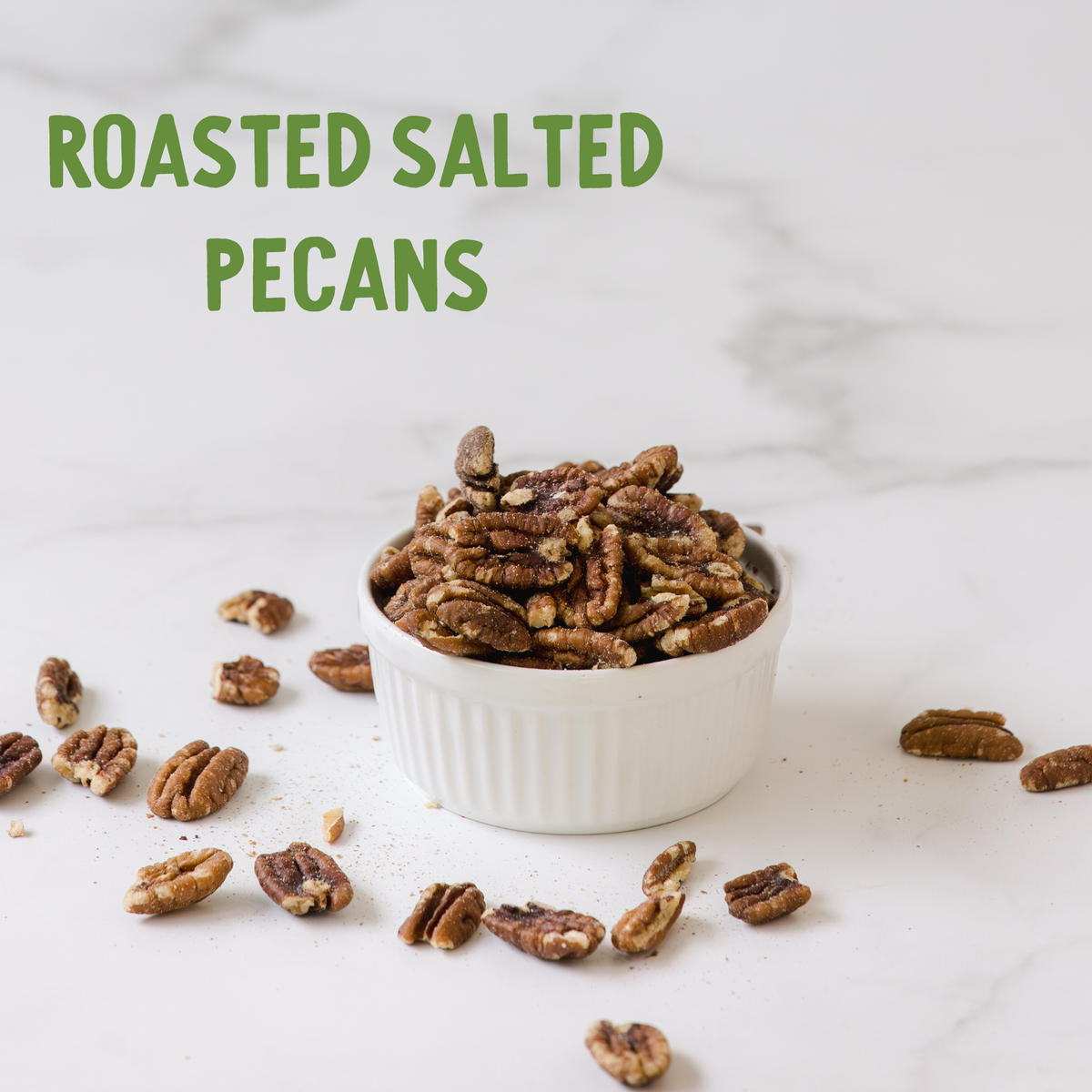 Roasted salted pecans in white ribbed ceramic bowl with scattered nuts on white marble surface