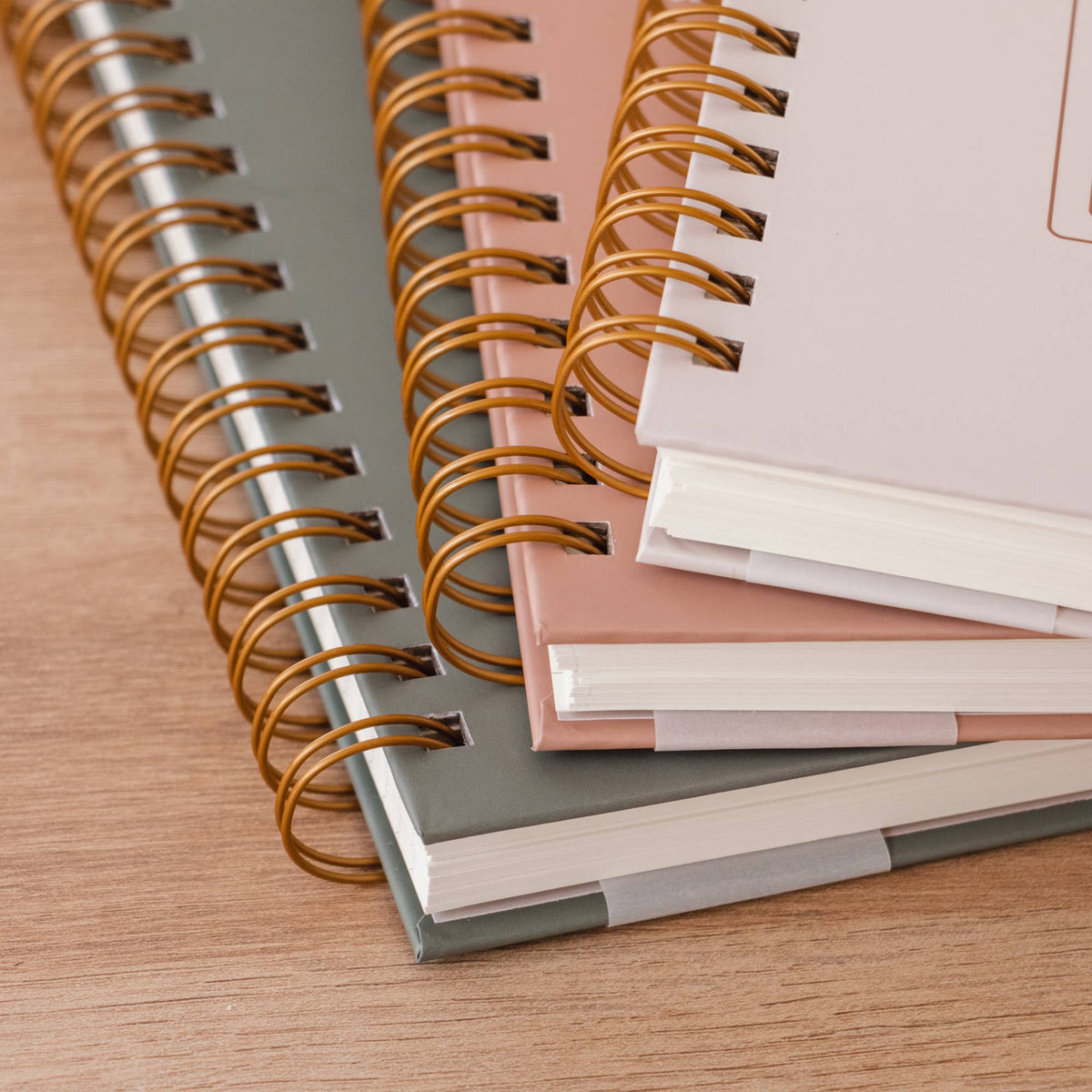 Three spiral-bound hardcover journals with gold metal bindings in pink, gray, and white covers, stacked on a wooden surface.