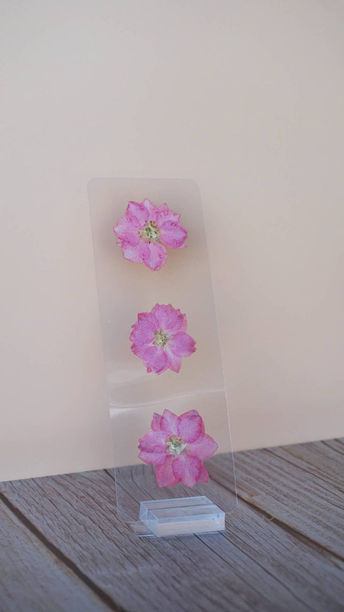 Three pressed magenta flowers with yellow stamens displayed on a clear acrylic bookmark stand against a light background