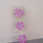 Three pressed magenta flowers with yellow stamens displayed on a clear acrylic bookmark stand against a light background