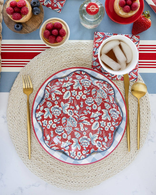 Red and white Floral Wave paper plate with botanical print, styled flatlay with gold cutlery, striped napkins, fresh berries, and drinks on marble surface.