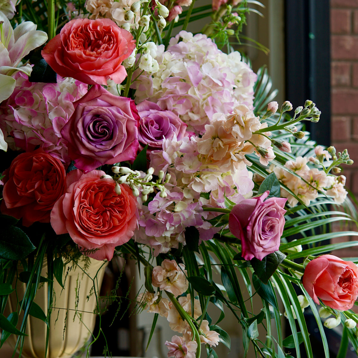 Funeral arrangement with coral and red peonies, burgundy roses, mauve hydrangeas, white stock flowers, and trailing green foliage in cream ceramic urn.