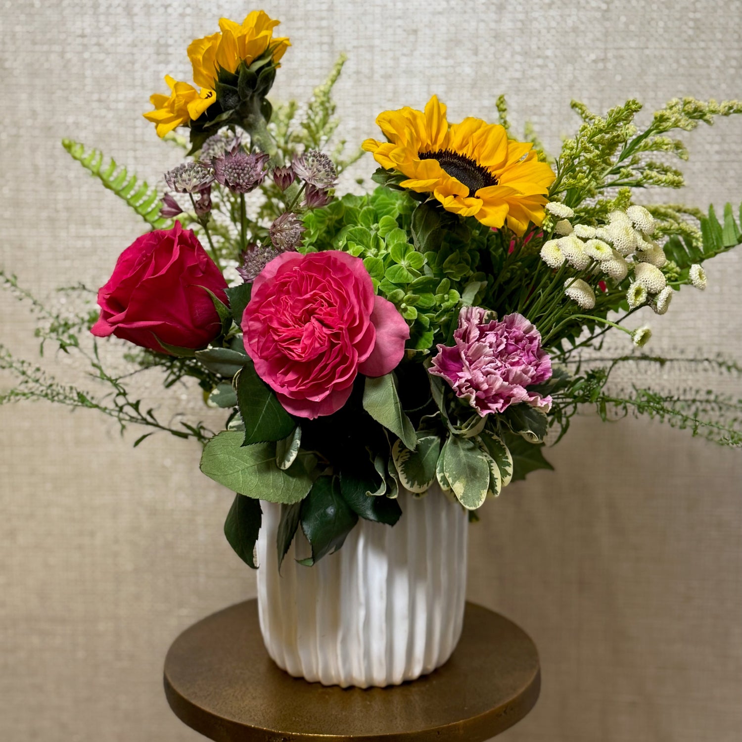 Vibrant flower arrangement in a white, textured vase. The arrangement features sunflowers, pink roses, a pink carnation, white button flowers, and various greenery, creating a cheerful and colorful display. The vase sits on a small, gold-colored table. The background is a neutral beige.