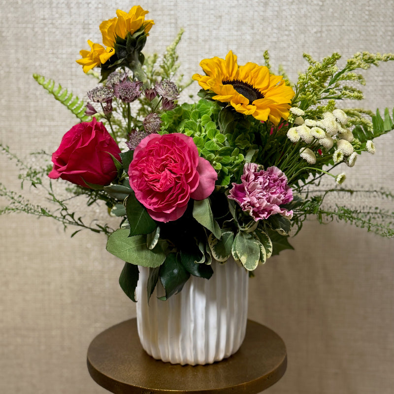 Vibrant flower arrangement in a white, textured vase. The arrangement features sunflowers, pink roses, a pink carnation, white button flowers, and various greenery, creating a cheerful and colorful display. The vase sits on a small, gold-colored table. The background is a neutral beige.