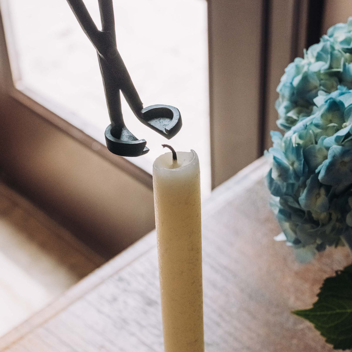 Black metal wick trimmer with curved blades positioned above a cream pillar candle on a windowsill, next to a blue-green succulent.