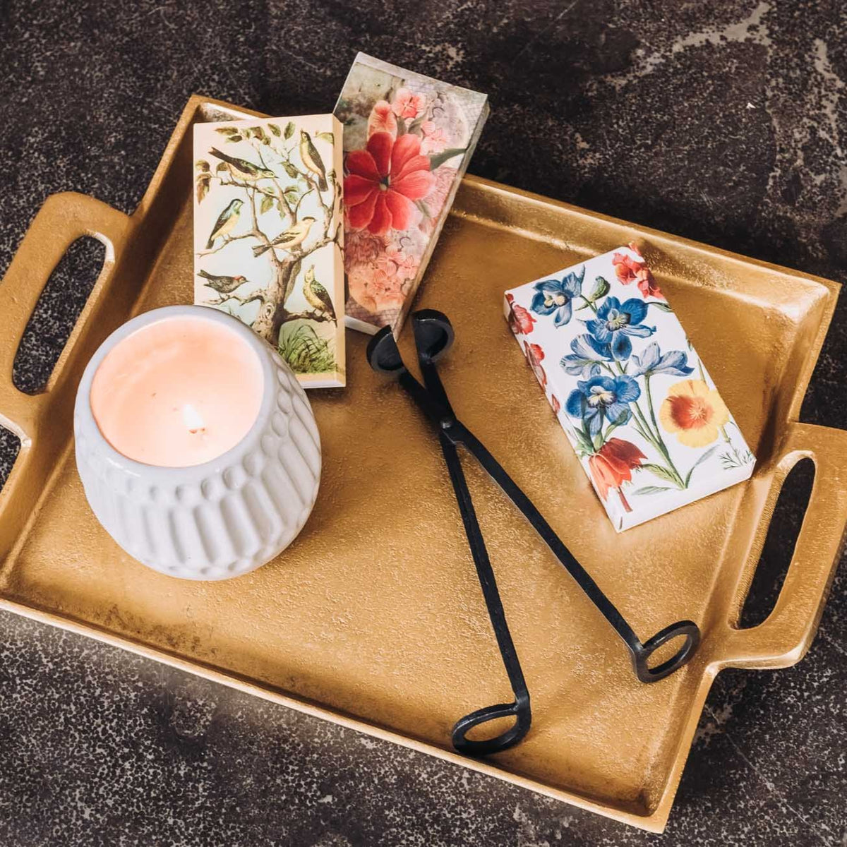 Black metal wick trimmer with curved handles displayed on a gold tray with a white candle, surrounded by botanical notecards