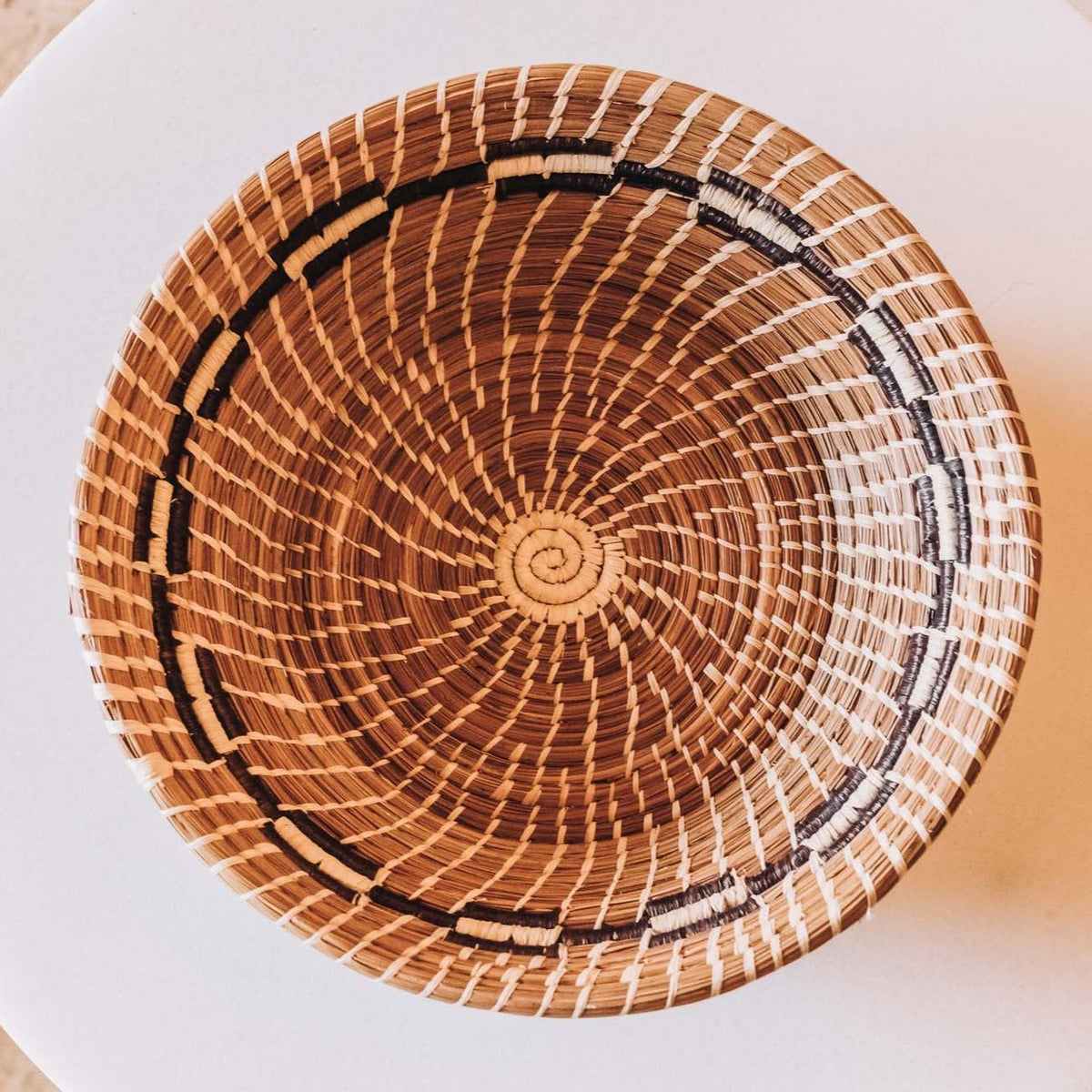 Handwoven round basket in natural tan and brown tones with white and black geometric accents, featuring a spiral center detail