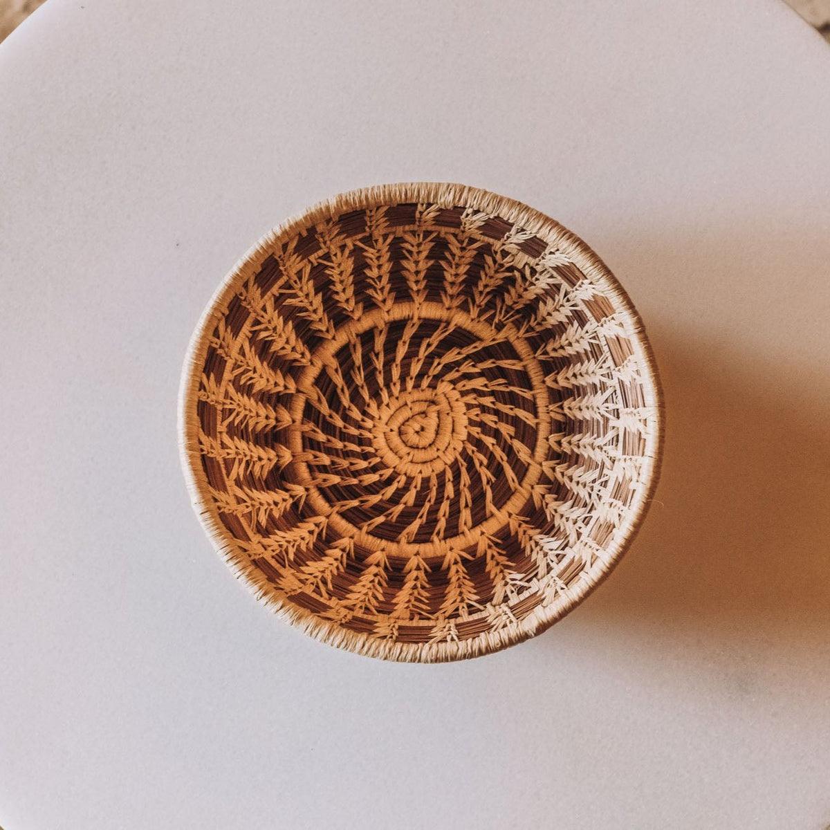 Round woven basket with spiral fern-leaf pattern in tan and brown, white rim, hand-woven from longleaf pine needles, flat lay on neutral background