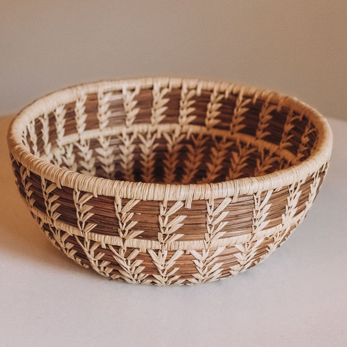 Round hand-woven basket bowl with geometric feather pattern in cream, beige, and brown pine needles