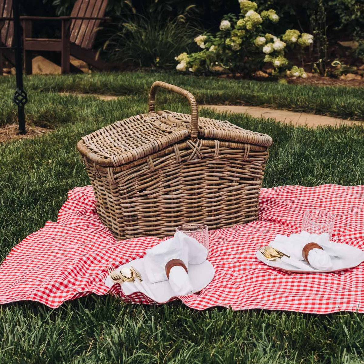 Handwoven rattan picnic basket in natural tan displayed on red and white gingham blanket in outdoor garden setting with place settings