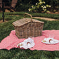 Handwoven rattan picnic basket in natural tan displayed on red and white gingham blanket in outdoor garden setting with place settings