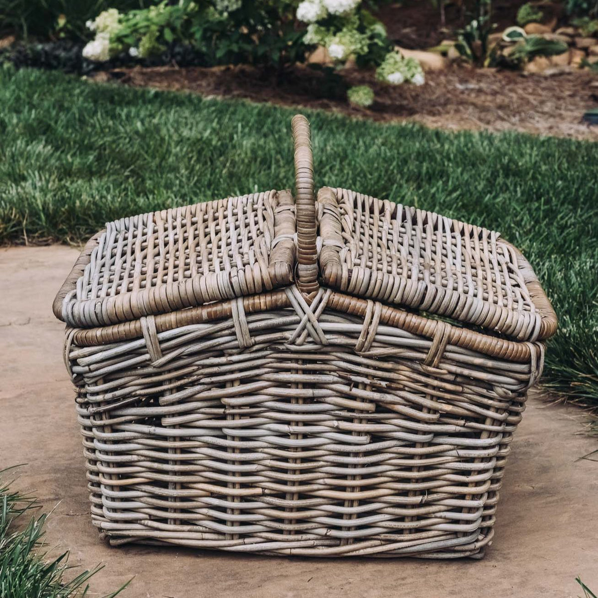 Whitewashed woven rattan picnic basket with dual handles and hinged lid, displayed on stone patio with garden background
