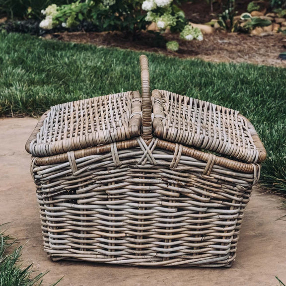 Whitewashed woven rattan picnic basket with dual handles and hinged lid, displayed on stone patio with garden background
