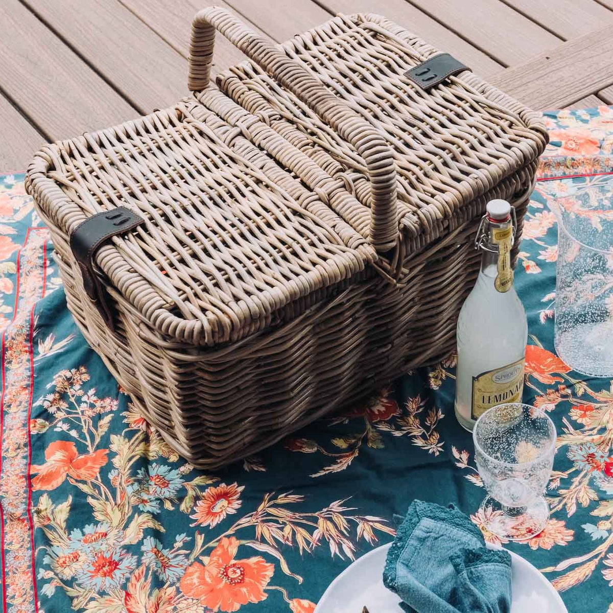 Natural rattan picnic basket with faux leather strap on patterned picnic blanket