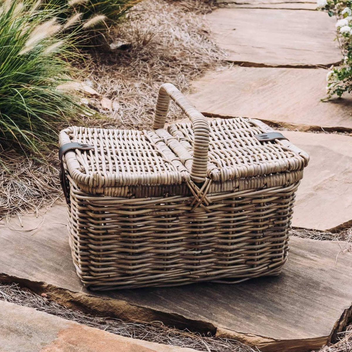 Brown rattan picnic basket with faux leather handle and padded interior, positioned on a stone pathway in a garden setting