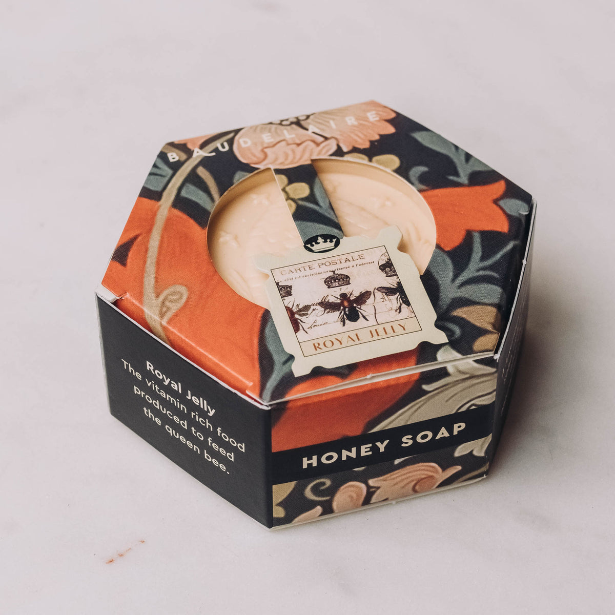 Cream-colored honey soap bar in hexagonal black and orange gift box with botanical design and bee motif, tied with cream ribbon