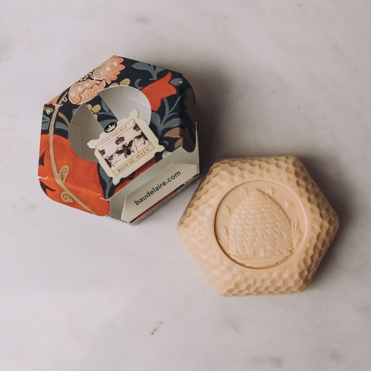 Cream-colored hexagonal honey soap bar with textured surface next to navy and coral floral gift box by Baudelaire