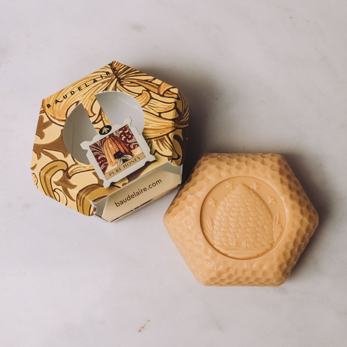 Cream-colored hexagonal bar soap with embossed honeycomb texture next to its botanical banana-patterned hex gift box by Baudelaire.