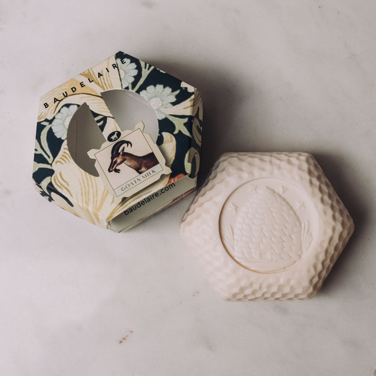Cream hexagonal bar soap with embossed details next to navy floral gift box labeled Baudelaire Goat's Milk