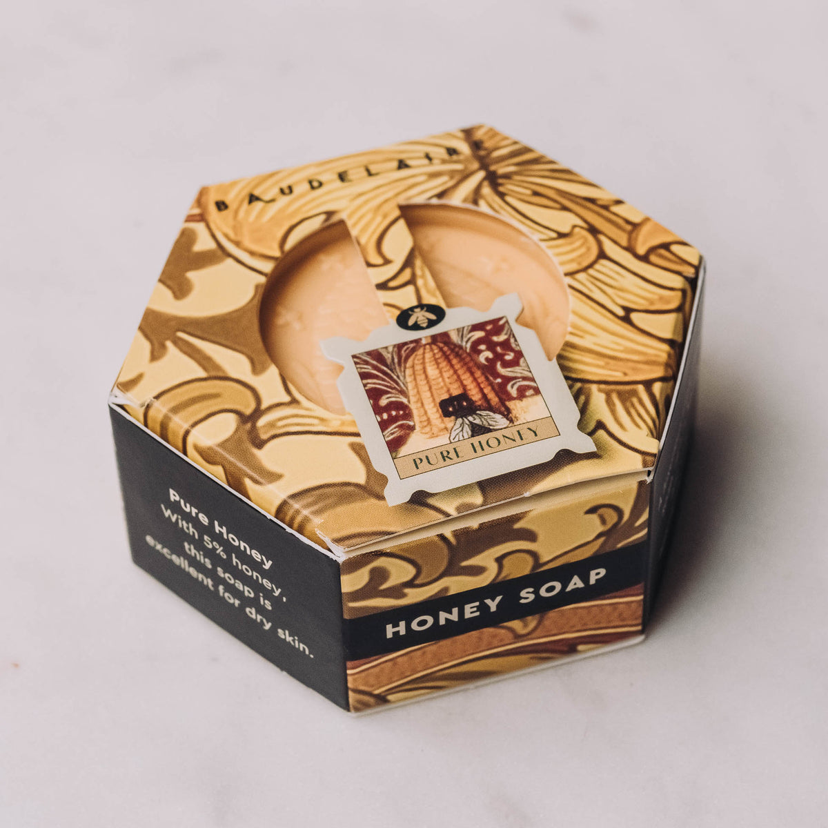 Hexagonal honey soap in cream-colored gift box with ornate botanical art, black band label, and pale honey-colored bar visible inside.